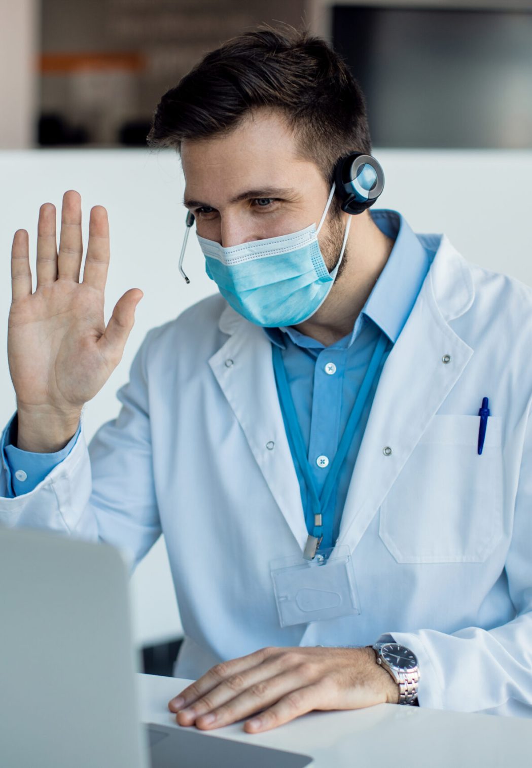 Doctor with face mask greeting someone while having video call over laptop in the hospital.